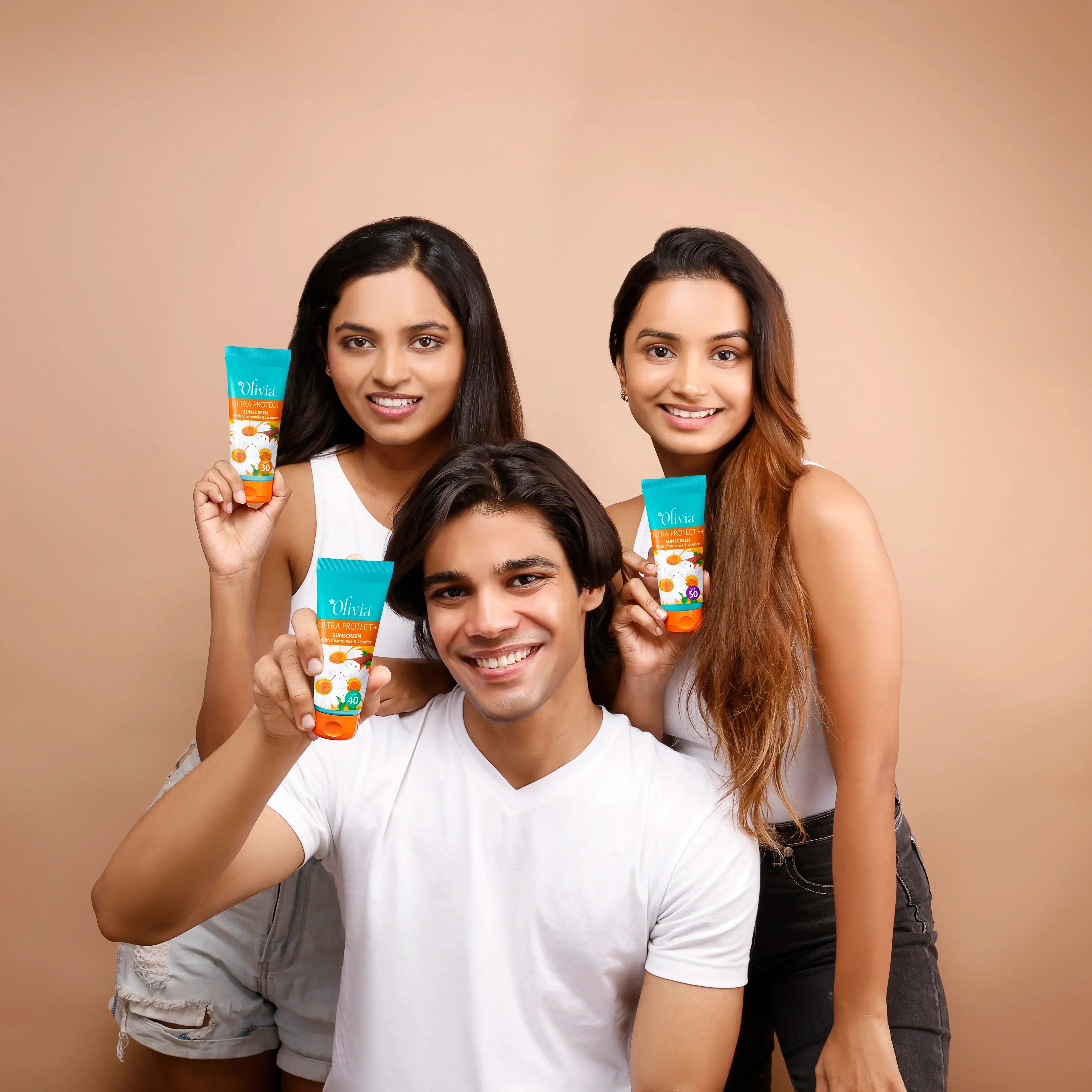 Three young Indian models smiling and holding Olivia Ultra Protect+ Sunscreen tubes in SPF 40 and SPF 50 variants with chamomile extract against a beige studio background